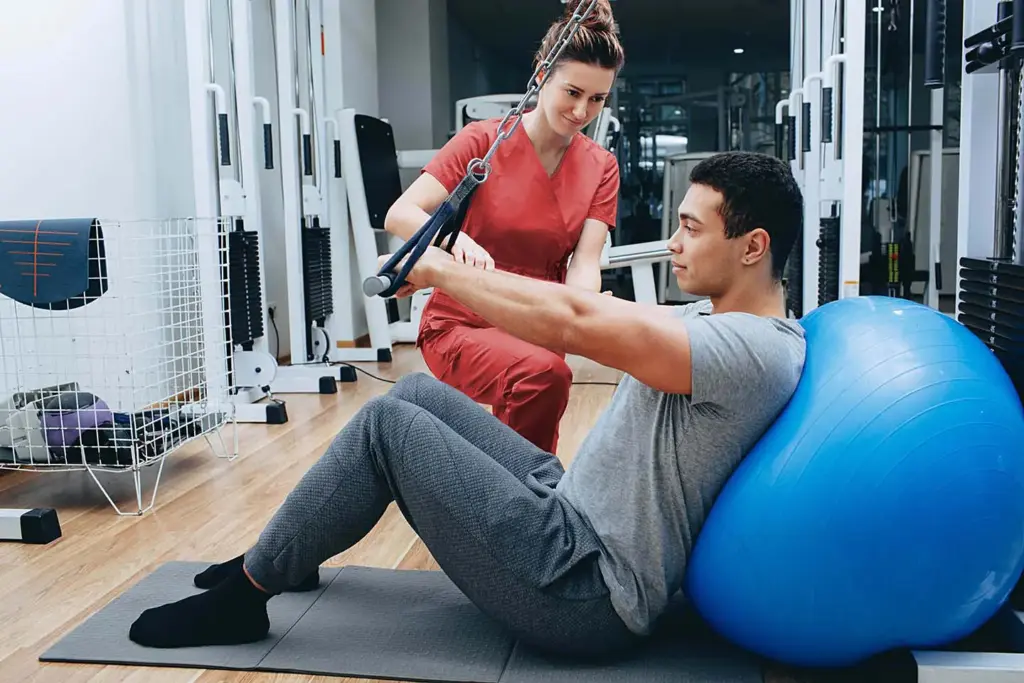 Physical therapist in red scrubs guides a man using a cable machine while leaning against a blue exercise ball.