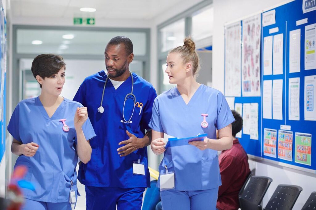 Three medical professionals in blue scrubs walking and talking in a hospital hallway.