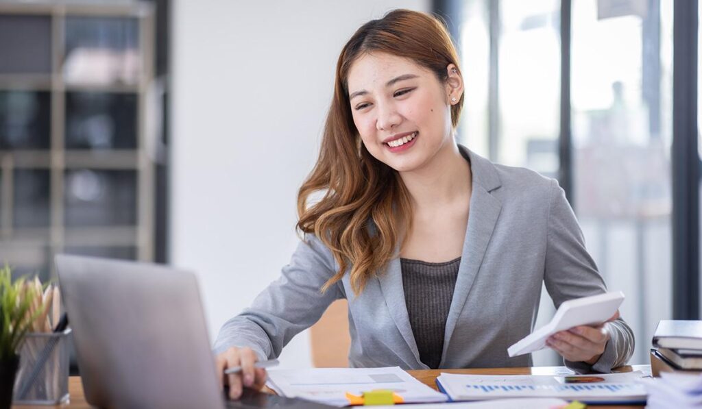 Smiling businesswoman in a grey blazer using a laptop and holding a calculator at her office desk.