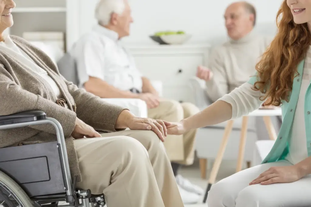 A caregiver holds the hand of an elderly woman in a wheelchair in a senior living facility.