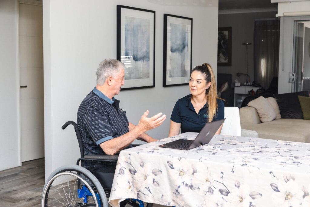 An elderly man in a wheelchair conversing with a woman at a table with a laptop.