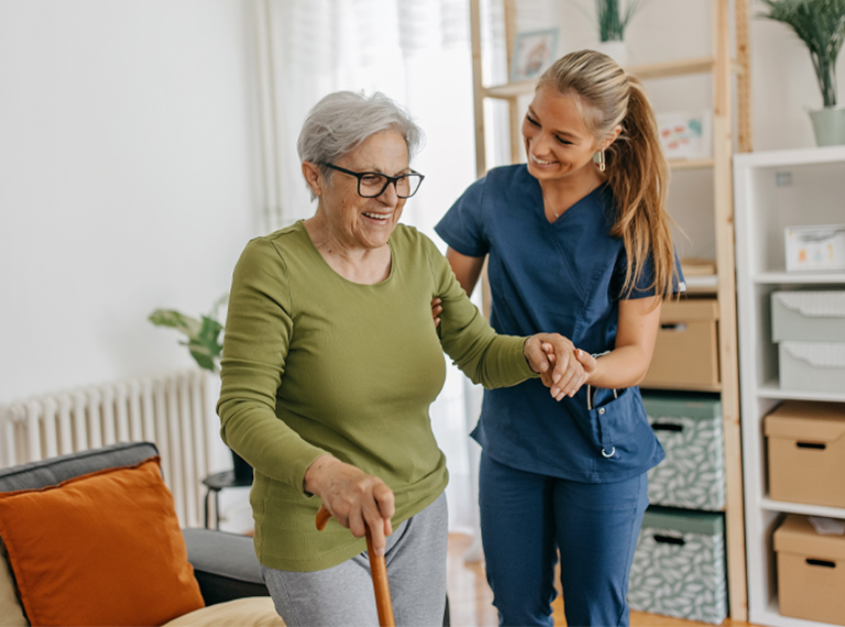 A smiling caregiver in blue scrubs assists an elderly woman using a walking cane.