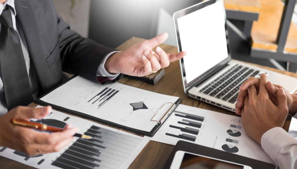 Business professionals reviewing financial charts and documents during a meeting at a desk with a laptop.