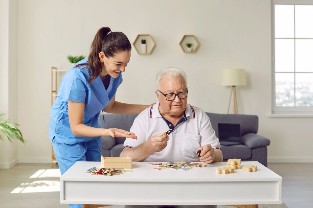 Smiling caregiver in blue scrubs assists an elderly man doing a cognitive puzzle game at a table.