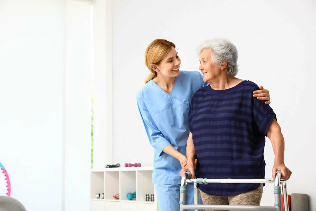 Smiling nurse in blue scrubs assisting an elderly woman using a walker.