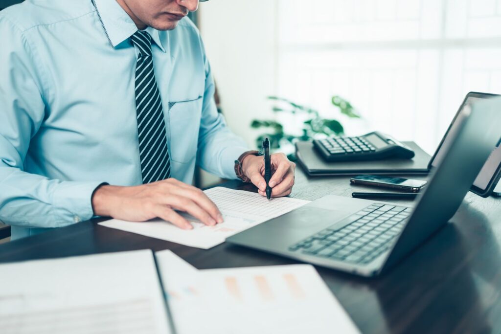 A professional in a dress shirt and tie reviewing and writing on financial documents at a desk with a laptop and calculator.