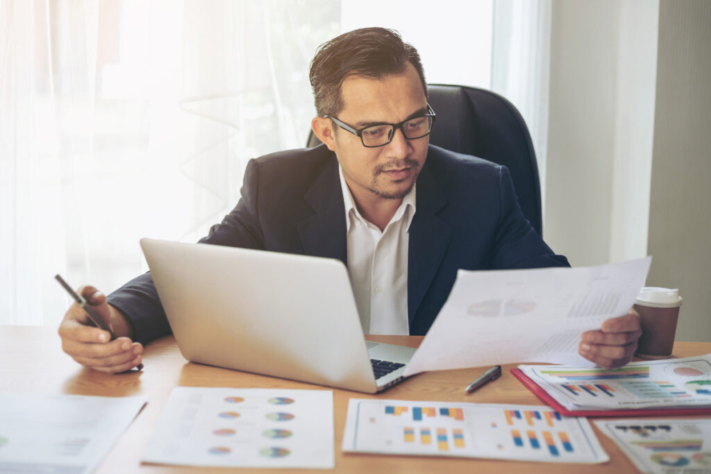Businessman wearing glasses sitting at a desk with a laptop, reviewing financial documents and charts.