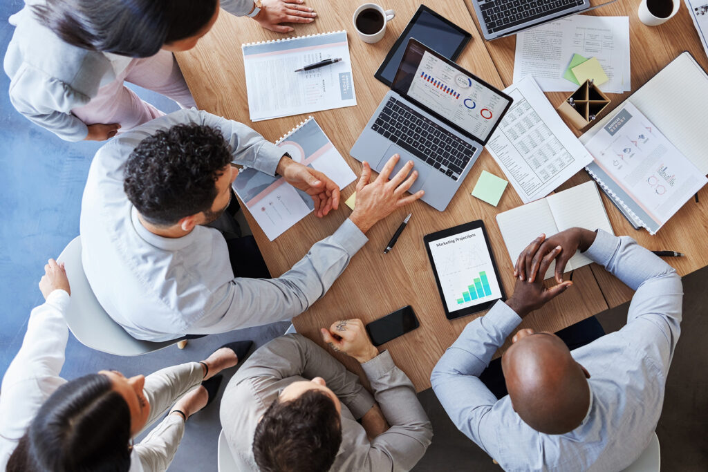 Overhead view of a diverse business team collaborating around a table with laptops, tablets, and data reports.