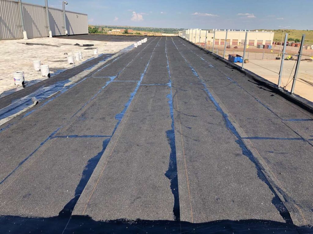 Black asphalt roofing membrane installed in long strips across a flat commercial rooftop under construction.