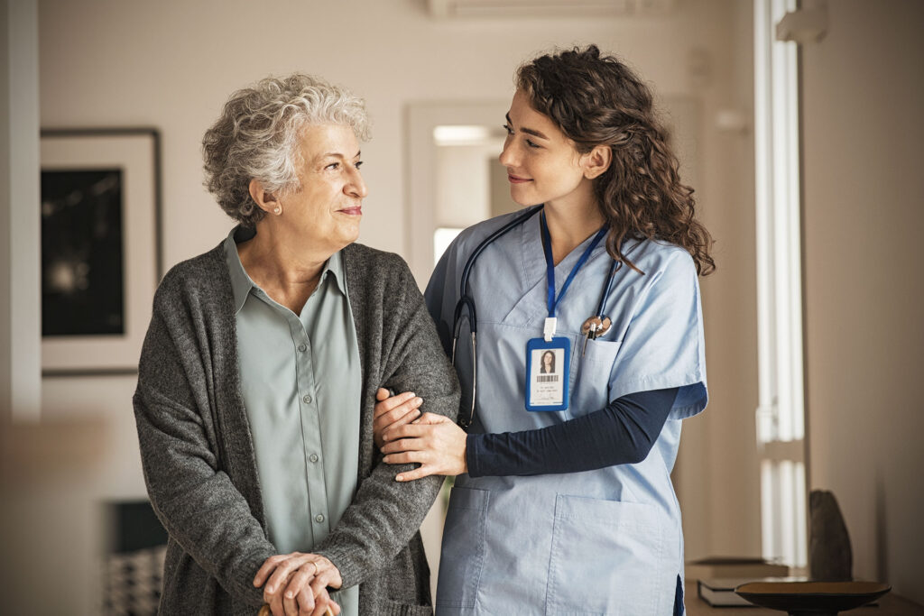 A female nurse in blue scrubs supports an elderly woman, both sharing a kind look.