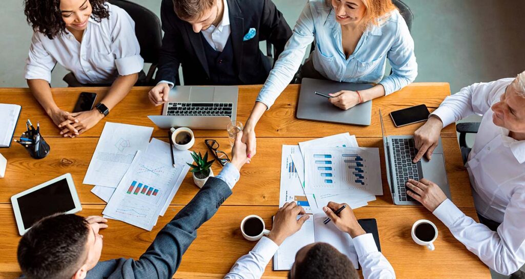 Overhead view of a diverse business team smiling as two colleagues shake hands across a wooden conference table.