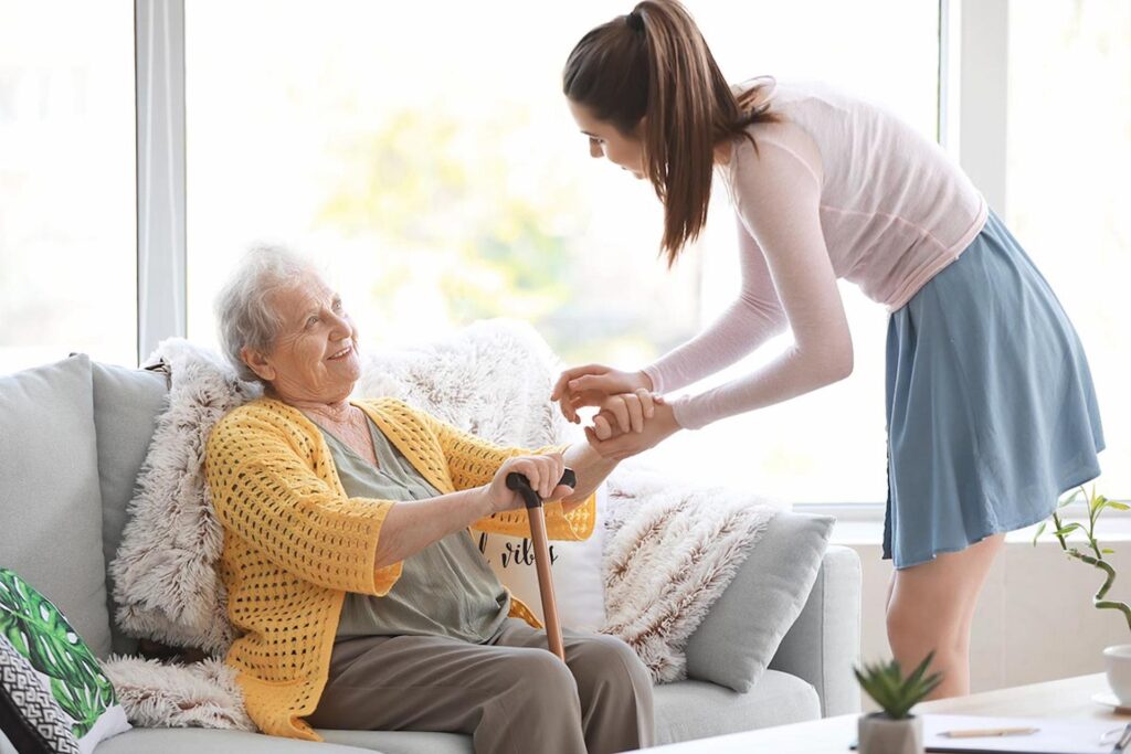 A young woman leans down to hold the hand of a smiling elderly woman sitting on a sofa.