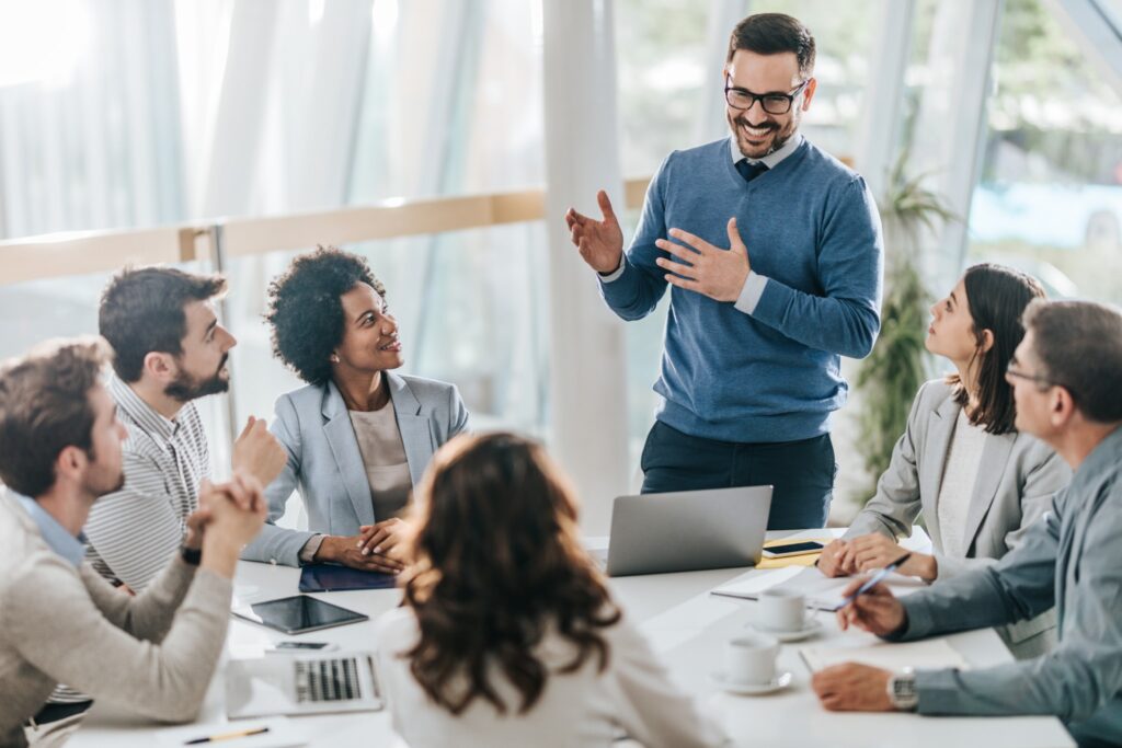 Smiling man in a blue sweater leading a meeting with a diverse group of colleagues around a conference table.