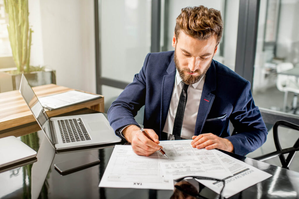 A man in a suit and tie working at a desk, filling out a tax form next to a laptop.