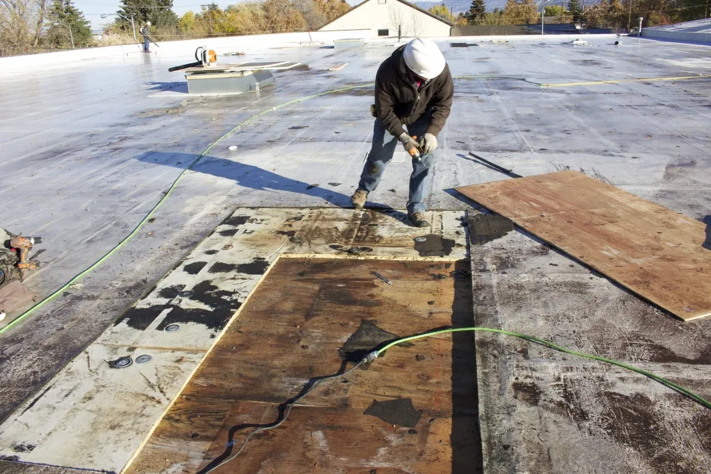 A roofer wearing a white hard hat uses a power drill while repairing a flat commercial roof.