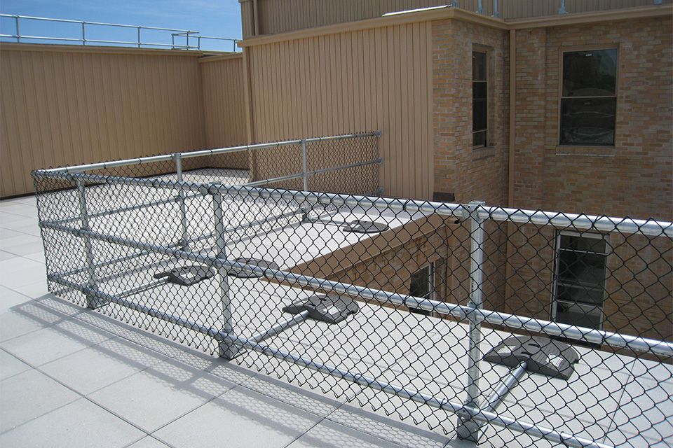 Chain-link safety fencing installed on a rooftop terrace next to a tan and brick building.