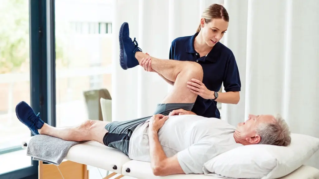 A female physical therapist assists an older man with a leg mobility exercise on a treatment table.