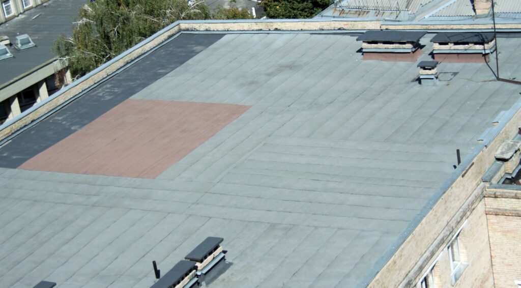 Aerial view of a flat grey rooftop featuring a rectangular reddish-brown patch and chimneys.
