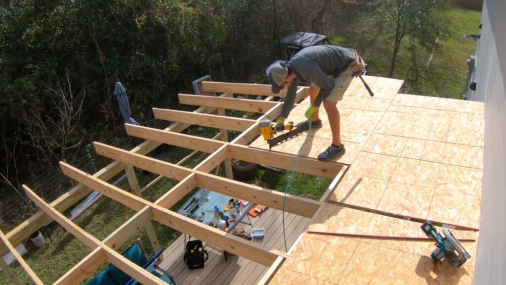 A construction worker installs OSB sheathing onto a wooden roof frame using a pneumatic nail gun.