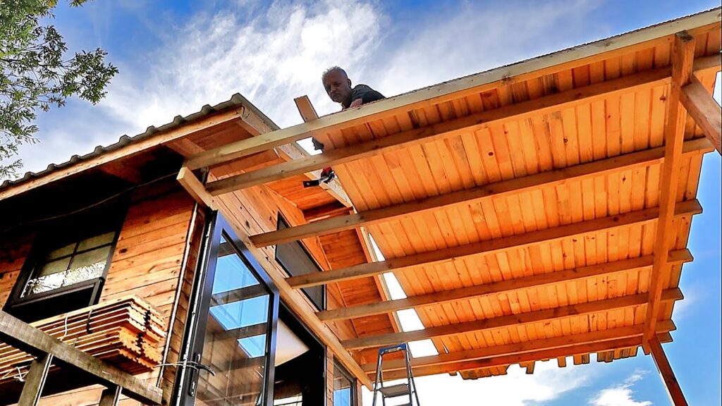 A man works on the wooden roof structure of a cabin under a blue sky.