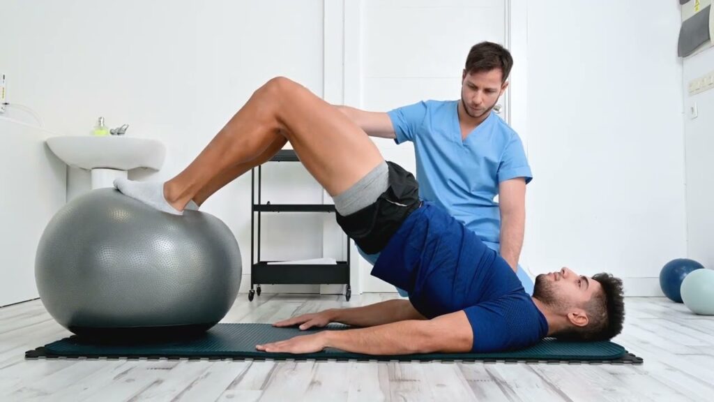 Physical therapist assisting a patient with a bridge exercise using a stability ball on a mat.
