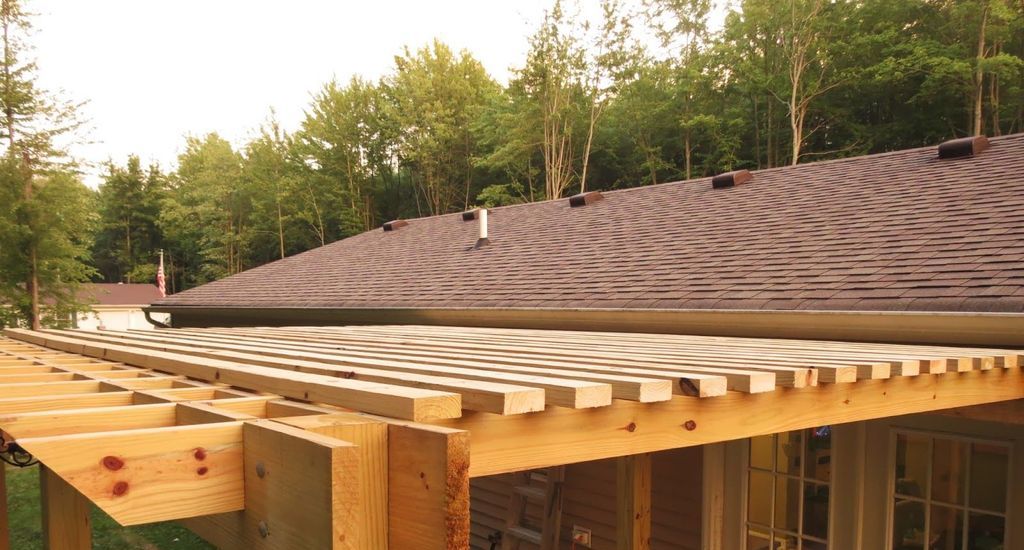 Wooden pergola frame under construction adjacent to a house with a brown shingled roof and background of green trees.