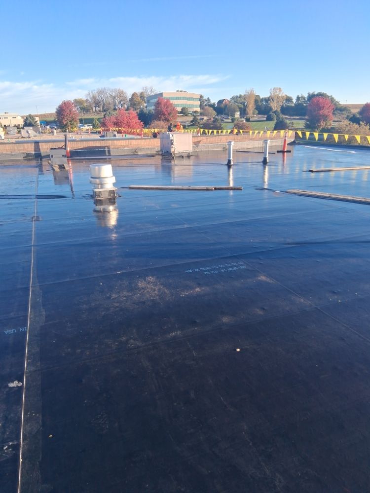 Wet black flat roof with ventilation pipes, construction equipment, and autumn trees in the distance.