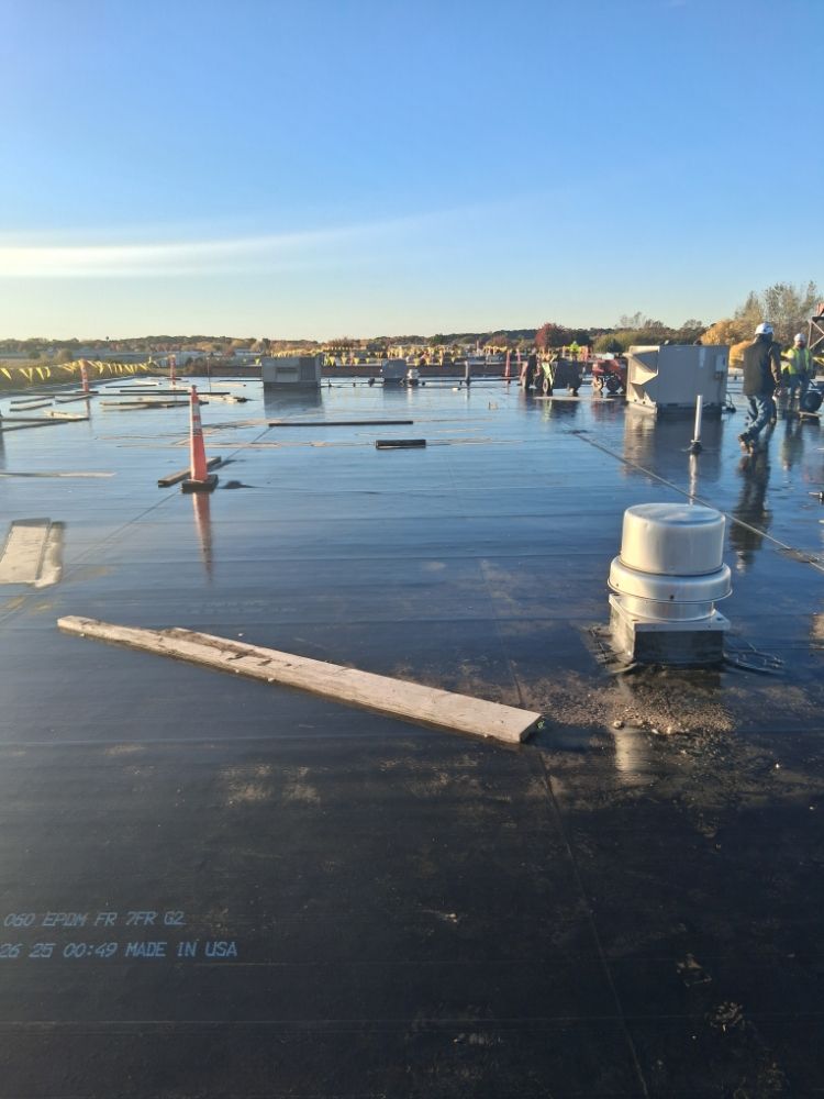 Workers on a large, flat black rubber roof under construction with HVAC units and safety cones.
