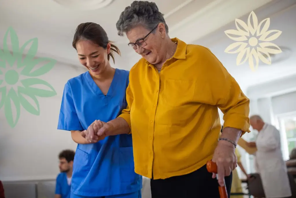 A nurse in blue scrubs smiling while helping an elderly woman in a yellow shirt walk with a cane.