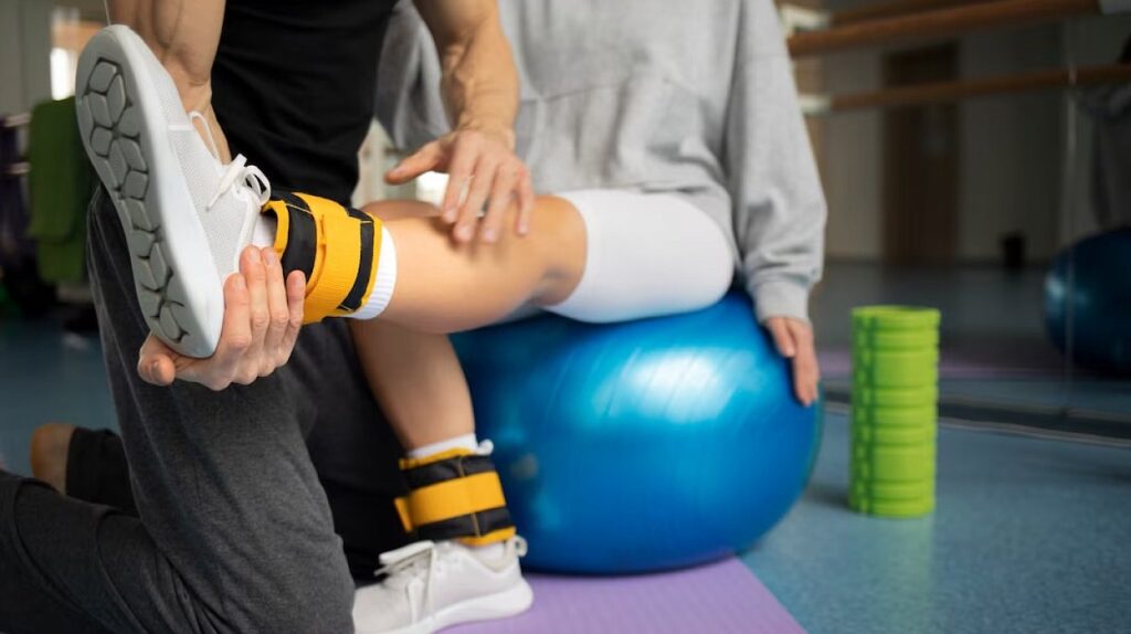 A physical therapist assists a patient performing leg exercises with weighted ankle straps while sitting on a blue stability