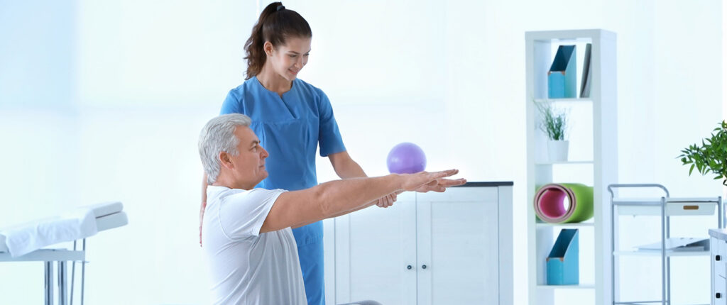 A physical therapist in blue scrubs assists an elderly man with arm stretching exercises in a bright clinic.
