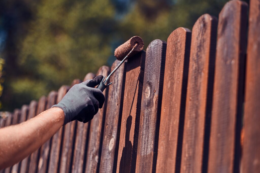 man in protective gloves is painting wooden fence in bright summer day