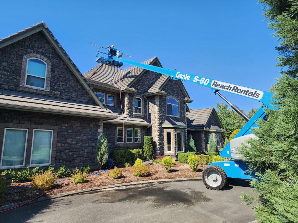 A blue boom lift extended to a worker on the roof of a large stone house.
