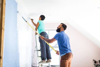 A man uses a long paint roller while a woman on a ladder paints a light blue wall in a bright room.