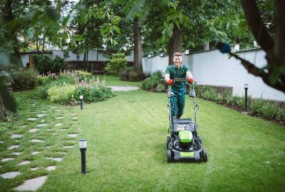 Smiling man in green uniform pushing a lawnmower across a lush green lawn in a garden.