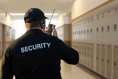 Security guard using a walkie-talkie in a school hallway lined with lockers.