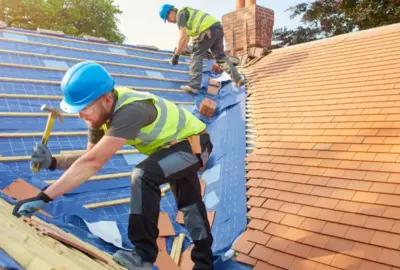 Two roofers in hard hats and safety vests installing tiles on a residential roof.