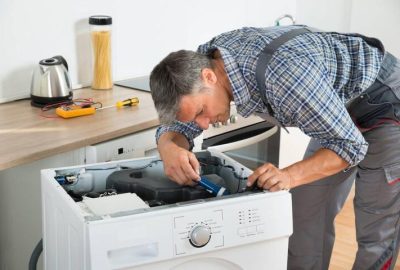 *   Image content: A man in work clothes (plaid shirt, grey overalls) is leaning over a white washing machine with its top pa