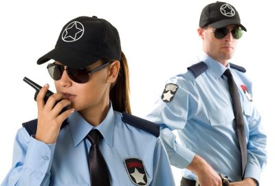 Two security guards in light blue uniforms, sunglasses, and black caps stand against a white background. One uses a walkie-ta