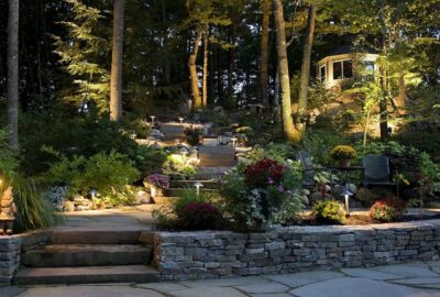 Evening view of a landscaped garden featuring stone steps, flower beds, path lighting, and a hilltop gazebo.