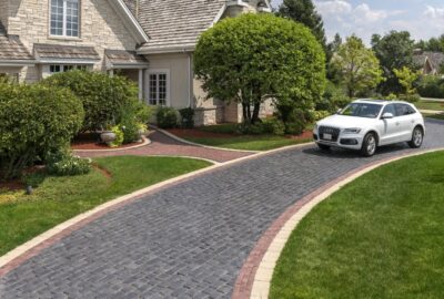 White Audi SUV parked on a patterned stone driveway leading to a large stone house with lush green landscaping.