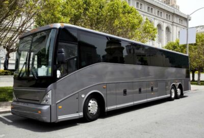 Modern silver luxury coach bus parked on a city street near a stone building and green trees.
