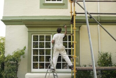 Painter in white overalls painting green trim around a window on a yellow house using a ladder and scaffolding.