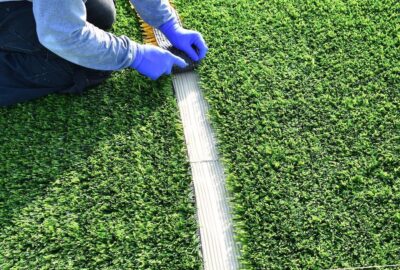 Worker in blue gloves installing artificial grass, pressing a seam joint with a tool.