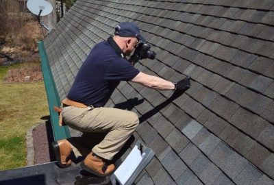 A roof inspector photographing shingles while crouching on a house roof.