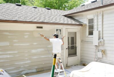 A man uses a paint sprayer to apply light beige paint to the horizontal siding of a white house.