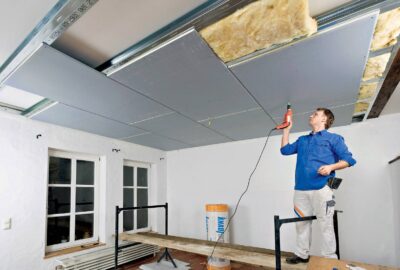 A man in a blue shirt uses a power drill to install drywall panels on a ceiling with exposed insulation.