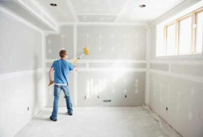 A man in a blue shirt and jeans uses a long-handled tool to sand or apply compound to drywall in a room under construction.