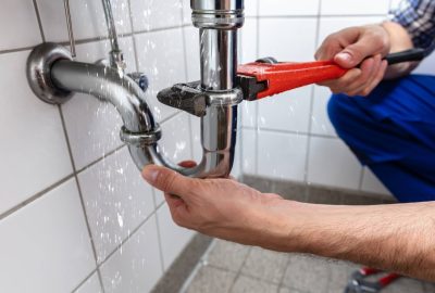*   Image content: A person (plumber) using a red pipe wrench to fix a leaking chrome P-trap under a sink. Water is spraying