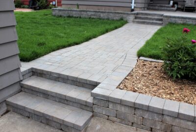Stone paver walkway and steps winding through a green lawn beside a mulched garden bed with pink peonies.